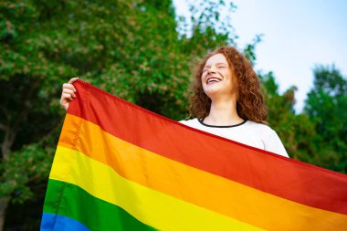 Lezbiyen mutlu bir kadının portresi, gülümseyen güzel bir kız, LGBT renginde bir gökkuşağı bayrağı tutan doğal bir arka planda güneşli bir yaz gününde. LGBT topluluğu, homoseksüel eşitlik kavramı