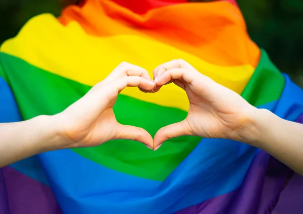LGBT kızı, lezbiyen kadın parmaklarıyla kalp şeklinde işaret gösteriyor, elleri gökkuşağı bayrağının arka planında. Homoseksüel aşk, ilişkiler.