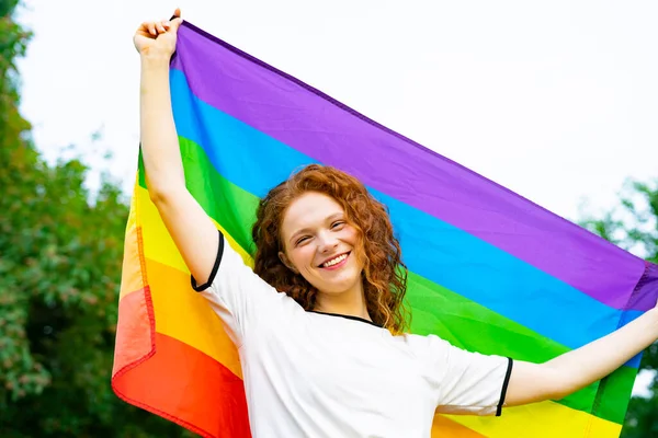 Lezbiyen mutlu bir kadının portresi, gülümseyen güzel bir kız, LGBT renginde bir gökkuşağı bayrağı tutan doğal bir arka planda güneşli bir yaz gününde. LGBT topluluğu, homoseksüel eşitlik kavramı