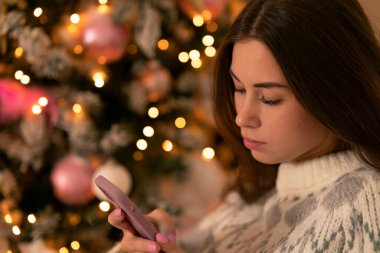 Neşeli bir Noel ağacının arka planında cep telefonu tutan üzgün kız. Akıllı telefonu olan kadın, kötü mesajlar, ayrılık mesajı, nahoş bir mesaj.