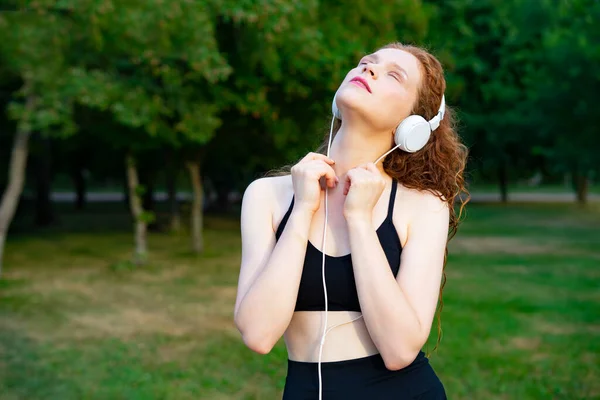 Mujer satisfecha y pensativa en ropa deportiva, auriculares escuchando música en el parque. Retrato de una mujer pelirroja —  Fotos de Stock