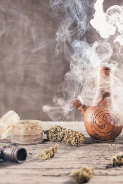 Ahşap yüzeyde tıbbi marihuana ile birlikte tüttüren eski bir kil nargilesi var. Çok duman var. Yakınlarda tıbbi esrar tomurcukları ve öğütücü var. film tahıl etkisi. Dikey yönelim