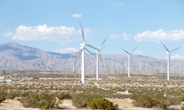 Yel değirmenleriyle çöl manzarası, arka planda dağlar olan vadide alçak büyüyen çalılar. Bulutlu mavi gökyüzü.
