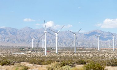 Yel değirmenleriyle çöl manzarası, arka planda dağlar olan vadide alçak büyüyen çalılar. Bulutlu mavi gökyüzü.