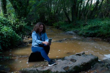 Kadın derenin kenarında oturmuş akıllı telefonuyla selfie çekiyor..