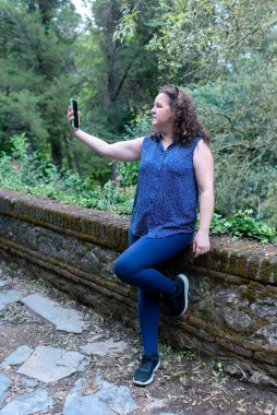 Kafkas bir kadının dikey fotoğrafı, biraz kıvırcık saçlı, ormanda akıllı telefonuyla selfie çekiyor.. .