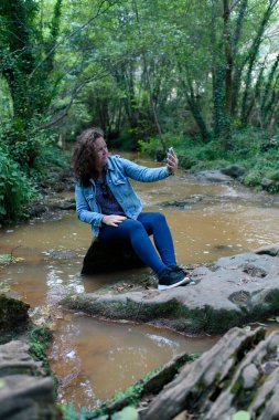 Dalgalı saçlı, beyaz bir kadının derenin yanında selfie çekerken çekilmiş dikey fotoğrafı..