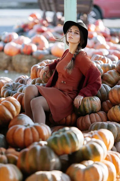 Stijlvolle vrouw zittend tussen een stapel rijpe oranje pompoenen op boerenmarkt in bruine trui, jurk en hoed. Gezellige herfst vibes Halloween, Thanksgiving dag — Stockfoto