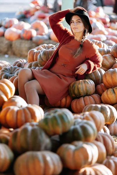 Stijlvolle vrouw zittend tussen een stapel rijpe oranje pompoenen op boerenmarkt in bruine trui, jurk en hoed. Gezellige herfst vibes Halloween, Thanksgiving dag — Stockfoto
