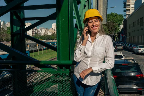 Beautiful young woman engineer or supervisor, wearing a safety helmet, talking on a cell phone while taking a short break inspecting a railroad pedestrian bridge. Concept of empowered woman, professional woman, professional job.