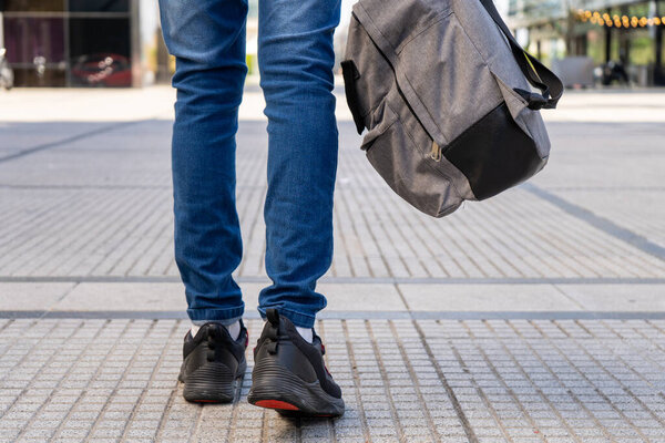 portrait of Legs of a young Latino walking down a street with an urban type backpack in his hand. He walks away from the camera.