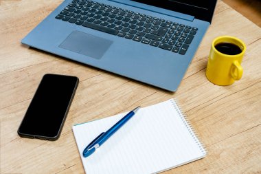 Boş bir not defterinin yanındaki dizüstü bilgisayar, çalışma masasındaki kahve ile birlikte kalem ve cep telefonu. Teknoloji, iletişim, ana ofis kavramı
