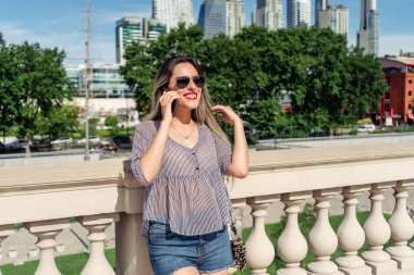 Güneşli bir günde arka planda ağaçlar ve binalarla cep telefonuyla konuşan güzel bir Latin kadın..