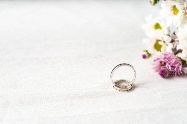 Beyaz bir yüzeyde, kenarında güzel beyaz ve mor çiçekler olan bir çift alyans. Bağlılık ve aşk kavramı. Boşluğu kopyala