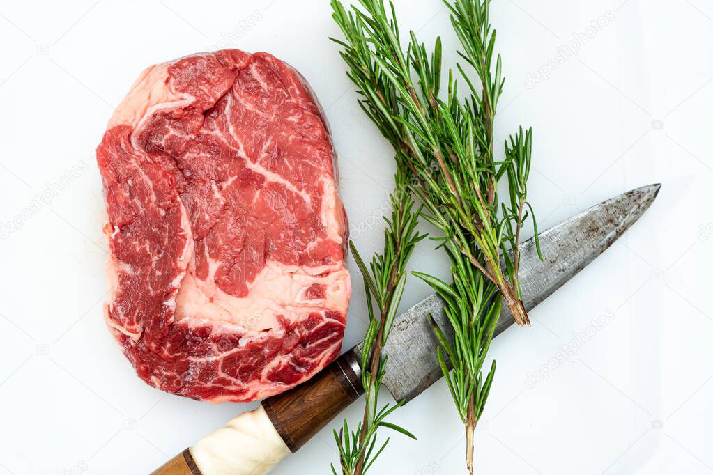 Close-up of a raw rib eye steak, one knife and a few sprigs of fresh rosemary on a white surface. Food concept. Overhead view.