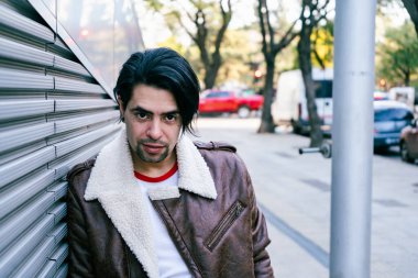 Close-up of a man in a leather jacket resting with his back against a modern wall of a building. Copy space