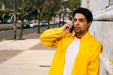 A portrait of a handsome latin young man in a yellow shirt talking on the phone in a street on a sunny day