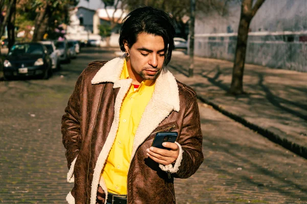 A handsome young Latino man in a leather jacket checking messages on his cell phone as he walks down a street.