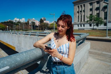 Genç kızıl saçlı kadın cep telefonundan mesajları kontrol ediyor, tavrı tatmin edici. Uzayı kopyala Peyzaj yönelimi.
