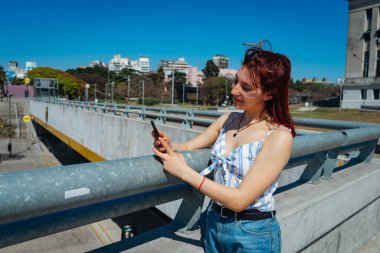 Genç, kızıl saçlı bir kadın cep telefonundan mesajları kontrol ediyor, tavrı tatmin edici. Uzayı kopyala Peyzaj yönelimi.