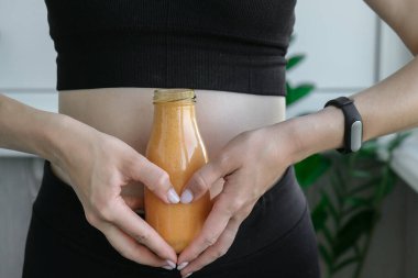 Kalp sembolü olarak, Smoothie içecek detoksunu zinde ve ince göbekli kadına tutuyordu. Fitness refah konsepti. Vücut bakımı, temiz yemek, kilo verme diyeti. Bağırsak veya üreme sorunu olan bir kadın. Evde antrenman yapıyorum. Göbek egzersizleri.