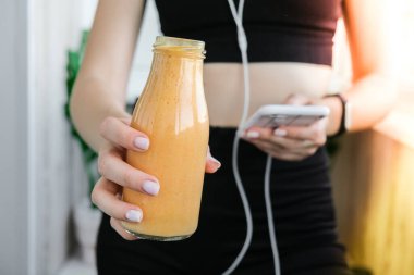 Genç bir kadın akıllı telefonu kontrol ediyor vejetaryen smoothie içiyor detoks içiyor. Evde yoga egzersizi yapan bir kadın. Çevrimiçi eğitim, temiz yemek, kilo kaybı, sağlıklı beslenme konsepti. Sağlıklı kalın. Zindelik
