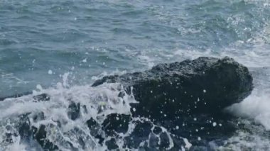 Dalgalar mavi okyanusta kayaların üzerinde kırılır. Okyanus dalgaları boş bir kaya kayasına çarpıyor. Beyaz köpükle kayalık kayalıklara çarpmak.