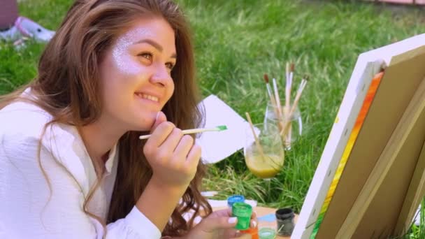 Молода приваблива жінка художник малює на природі. Художники палітрують фарбами та пензлем хоббі. Творчий художник малює на полотні під відкритим небом. Візуальне мистецтво на зеленій траві в міському парку. Повільний рух — стокове відео