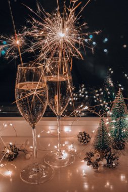 İki bardak köpüklü şampanya ve maytap. Sarı ışık saçan koyu bir arka plan. Masada Noel ağacı oyuncakları var. Yeni yılın Bengal ateşi. Noel kış tatili