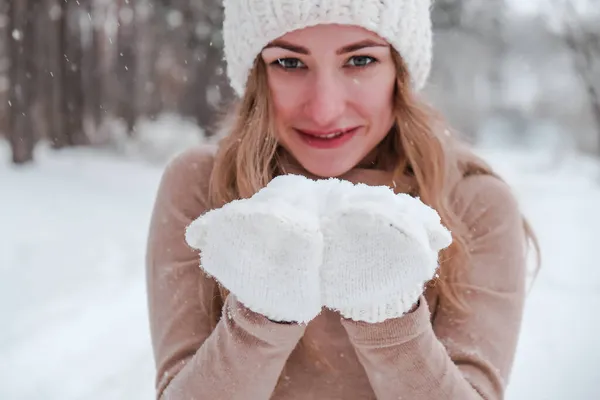 Noël, vacances et concept de saison. Jeune femme heureuse soufflant de la neige dans la forêt d'hiver nature. Vêtements chauds gants tricotés et chapeau. — Photo