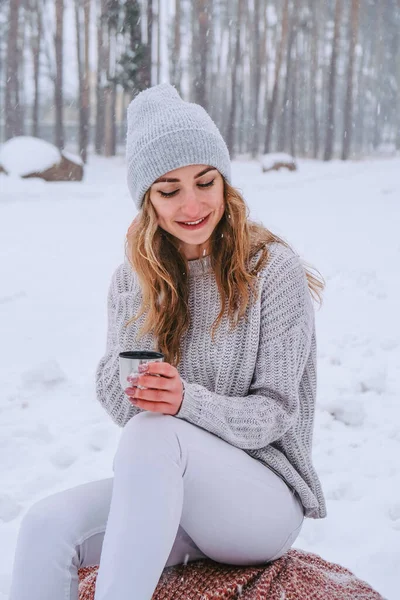 Cute Caucasian girl in a white sweater and hat in a snowy forest Park drinking hot tea or chocolate from a thermos. Girl Wearing Warm Winter Clothes — Stock Photo, Image