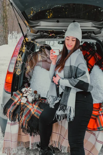 Brunette woman wrapped in blanket in trunk car drinking coffee tea from thermos. Travel in winter. Car decorated with festive Christmas lights. Outdoor picnic — Stock Photo, Image