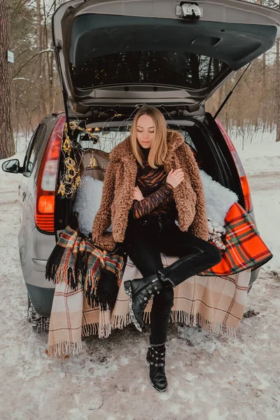 Mujer rubia envuelta en manta en el coche del maletero. Viajar en invierno. Coche decorado con luces navideñas festivas. Picnic al aire libre — Foto de Stock