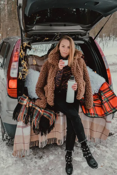 트렁크에 담요로 둘러 싸인 금발의 여자가 열모에서 커피를 마시고 있습니다. 겨울 여행. 크리스마스 조명으로 장식된 차. 옥외 소풍 — 스톡 사진