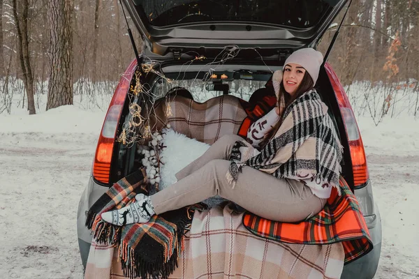 Bagaja sarılmış esmer kadın termostan kahve içiyor. Kışın seyahat. Noel ışıklarıyla süslenmiş bir araba. Açık hava pikniği — Stok fotoğraf