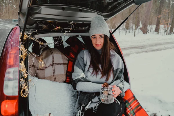 Mujer morena envuelta en manta en el coche del maletero. Viajar en invierno. Coche decorado con luces navideñas festivas. Picnic al aire libre — Foto de Stock