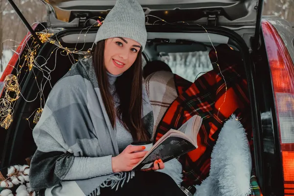 Brunette woman wrapped in blanket in trunk car drinking coffee tea from thermos. Travel in winter. Car decorated with festive Christmas lights. Outdoor picnic — Stock Photo, Image