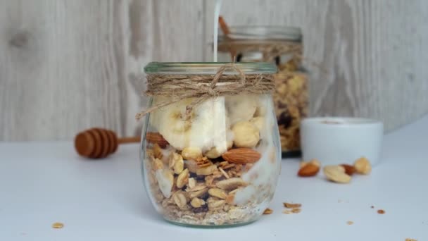 Sarapan sehat 4k. Oatmeal Granola dengan yogurt hijau menuangkan dan kacang pisang muesli dalam toples pada latar belakang cahaya. Konsep diet vegan, vegetarian dan penurunan berat badan. Menu detox. — Stok Video