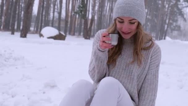 Nettes kaukasisches Mädchen in weißem Pullover und Hut in einem verschneiten Waldpark, das heißen Tee oder Schokolade aus einer Thermoskanne trinkt. Mädchen tragen warme Winterkleidung — Stockvideo