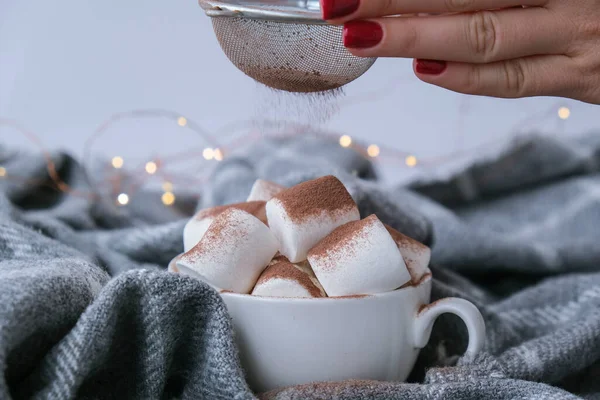 Adding cacao powder on top of hot chocolate with marshmallows in white cup on sweater background. Cozy christmas lights. Preparation for new year holidays atmosphere