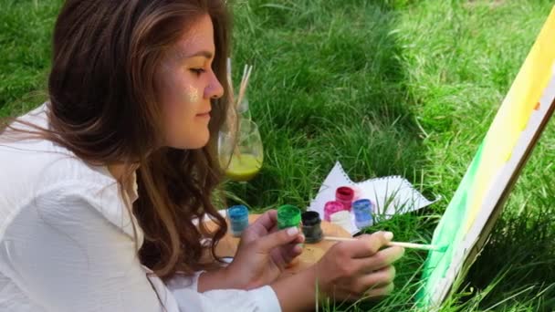 Νεαρή ελκυστική γυναίκα καλλιτέχνης ζωγραφίζει στη φύση. Παλέτα καλλιτεχνών με χρώματα και πινέλο Hobby. Δημιουργική ζωγραφική καλλιτέχνη σε καμβά στην ύπαιθρο. Εικαστική τέχνη στο πράσινο γρασίδι στο πάρκο της πόλης. Αργή κίνηση — Αρχείο Βίντεο