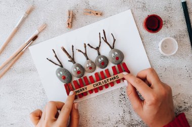 Adım adım. Üst Manzara. İşlemci çocuk işi. Kendin yap Mutlu Noeller geyiği çakıl taşlarından, deniz taşlarından, mandallardan ve beyaz arka planda dallardan oluşan kartpostalda. Doğal Eko tarzı diy. Hediye fikri..