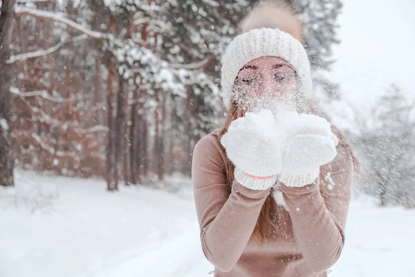 Noel, tatil ve sezon konsepti. Genç mutlu kadın kış ormanlarında kar üflüyor. Sıcak giysi, eldiven ve şapka.. — Stok fotoğraf