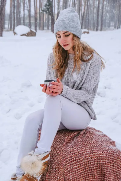 Мила кавказька дівчина в білому светрі і капелюсі в сніжному лісовому парку п'є гарячий чай або шоколад з термоса. Дівчинка одягає теплий зимовий одяг — стокове фото