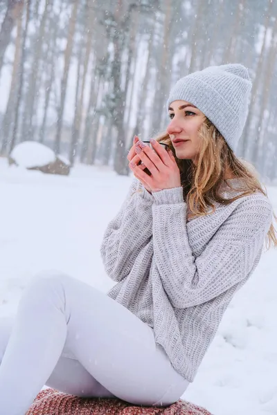 Cute Caucasian girl in a white sweater and hat in a snowy forest Park drinking hot tea or chocolate from a thermos. Girl Wearing Warm Winter Clothes — Stock Photo, Image