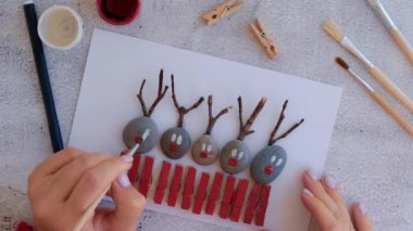 Çakıl taşları, deniz taşları, çamaşır mandalları ve beyaz arka planda dallardan oluşan kartpostalda Mutlu Noeller geyiği yapmak. Doğal Eko tarzı diy. Hediye fikri. Adım adım. Üst görünüm.