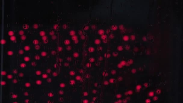 Dešťové kapky na okenní sklo zblízka s červeným světlem. Noční odraz světla na mokrém skle. Textura pozadí kapičky — Stock video