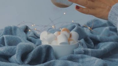 Sıcak çikolatanın üstüne kakao tozu ekliyorum. Süveter arka planında beyaz bardakta marşmelov. Sıcak Noel ışıkları.