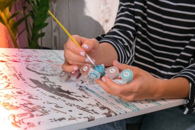 Tabloya numaralarla ve numaralandırılmış boya kavanozlarıyla resim yapmak. Kadın fırçayla resim çiziyor. Yaratıcı hobi. Yeni başlayanlar için resim yapmak. Evde kilitli kalmak için boş zaman aktivitesi, stres önleyici bir fikir. Meditasyon, rahatla.