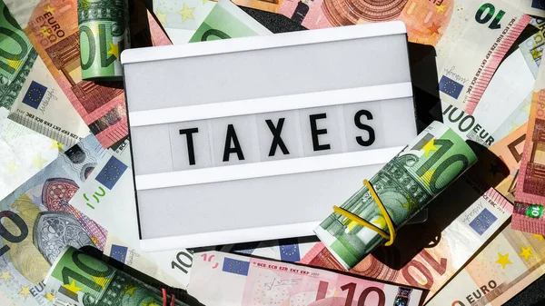 Tablica z napisem TAXES czarnymi literami wokół banknotów euro. Koncepcja płacenia podatków i ich wypełniania. Pieniądze, Biznes, finanse, inwestycje, oszczędności i korupcja — Zdjęcie stockowe